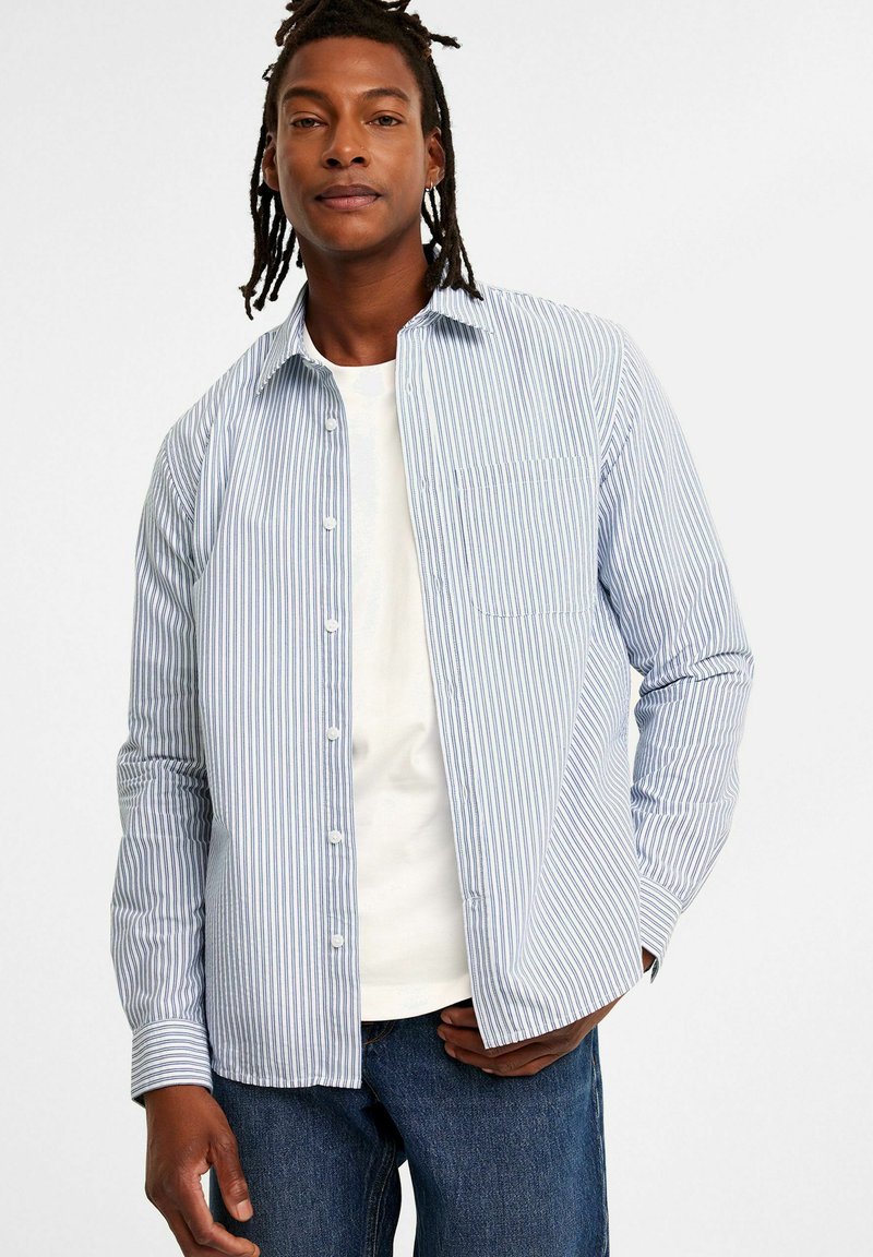 Blauw-wit gestreepte button-up shirt met kraag, lange mouwen en een borstzak, gedragen over een witte T-shirt en blauwe spijkerbroek.