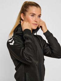 Noor naine blondide hobusesabadega kohandab oma musta lukuga dressipluusi kapuutsi, millel on valge noolelogoga õlal.