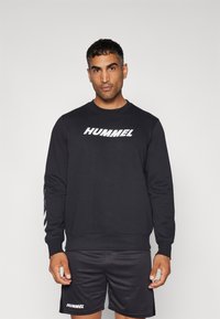 Sort sweatshirt med hvid "HUMMEL" logo på fronten, lange ærmer og ribbede manchetter. Bæres med matchende sorte shorts.