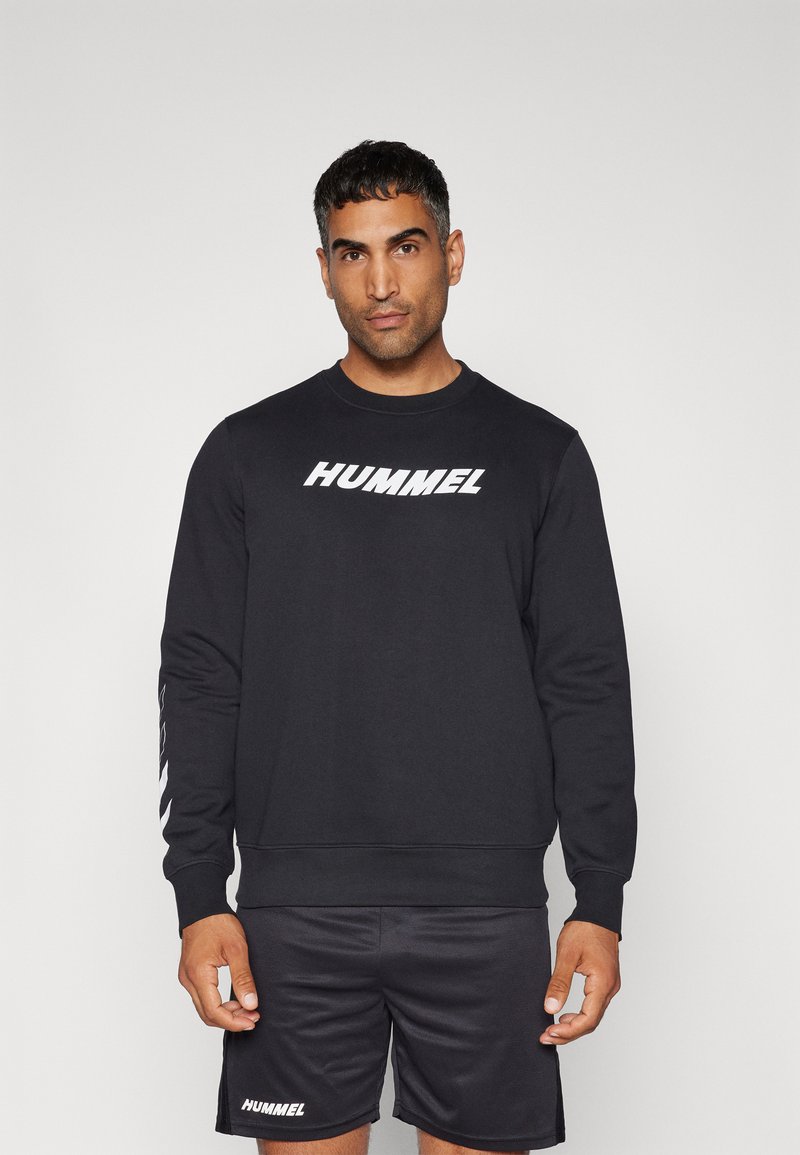Sort sweatshirt med hvid "HUMMEL" logo på fronten, lange ærmer og ribbede manchetter. Bæres med matchende sorte shorts.