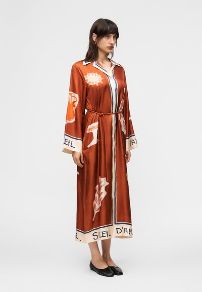 Γυναίκα που φοράει ένα μακρύ φόρεμα σε σκουρό πορτοκαλί με κουμπιά, με τυπωμένα σχέδια, ζώνη στη μέση και μαύρα επίπεδα παπούτσια, στέκεται μπροστά σε απλό λευκό φόντο.