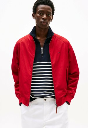 Rode bomberjack met een ritssluiting aan de voorkant, geribde manchetten en een marineblauwe gestreepte blouse eronder. Witte broek met zichtbare ritssluiting.
