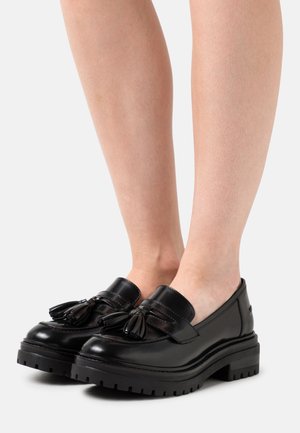 Mocassins en cuir noir avec une semelle en caoutchouc épaisse et des détails en pampilles. Texture lisse, bout rond et design minimaliste.