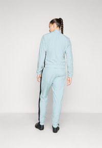 Costum sport din material albastru deschis, cu un design ajustat; include manșete elastice, dungi negre pe lateral și pantaloni de tip jogger asortați cu ajustări la glezne.