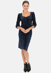 Granatowa sukienka z welwetu, z kwadratowym dekoltem, długimi bufkami, detalami marszczenia i dopasowanym krojem, w komplecie z czarnymi botkami za kostkę.