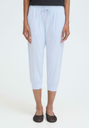 Pantalon court bleu clair avec taille élastique, cordon de serrage et ourlets retroussés, en tissu doux. Chaussures plates noires portées.