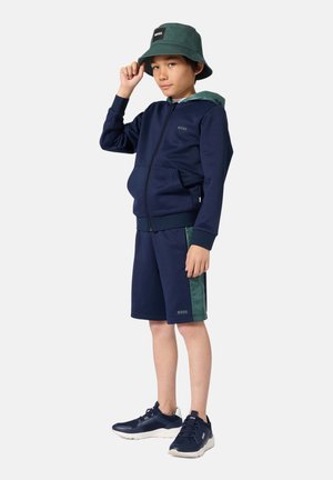 Marineblauwe hoodie met rits en groene accenten, bijpassende korte broek en marineblauwe sneakers. Het model draagt een groene bucket hat met een logo.