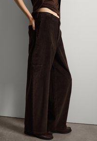 Pantaloni a gamba larga in corduroy marrone con dettagli delle cuciture visibili e tasche laterali, abbinati a calzature scure su una superficie texture.