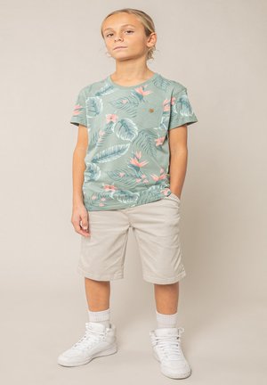 Enfant debout les mains dans les poches, portant un t-shirt vert à fleurs, un short beige, des chaussettes blanches et des baskets blanches montantes.