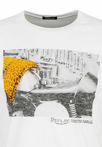 Camiseta de algodón blanca con un estampado gráfico de una motocicleta y herramientas, acentuada por un detalle de estampado de leopardo amarillo. Presente el logo de la marca.