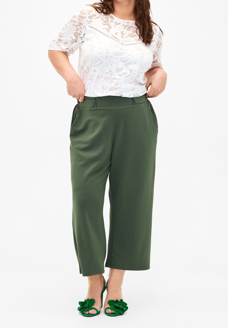 Blouse en dentelle blanche à motif floral, pantalon vert à jambes larges avec poches latérales et fermeture par bouton, sandales vertes à talons avec détail fleuri.