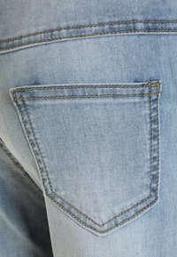 Jeans de denim azul claro con un bolsillo trasero, que presentan una textura sutil y un hilo dorado en contraste a lo largo de las costuras y los bordes del bolsillo.