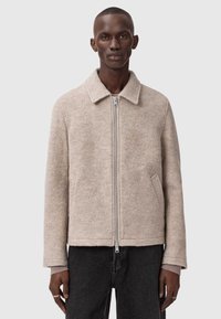 Beige uldjakke med flad krave, lynlås foran, to sidelommer og en struktureret overflade. Båret over mørke jeans.