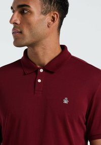 Bordeaux polo shirt met een kraag, voorzien van twee witte knopen en een klein geborduurd logo van een pinguïn op de linkerkant van de borst. Glad stoftextuur.