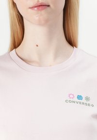 Šviesiai rožinė medvilninė marškinėliai su raukšlėtu apykaklės kraštu ir mažais logotipo dizainais su spalvingais akcentais bei žodžiu "CONVERSE" žaliai.