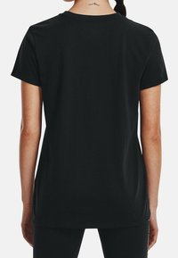 Vrouw van achteren gezien, gekleed in een effen zwarte T-shirt met korte mouwen en zwarte broek, met een kleine golftattoo op de nek.