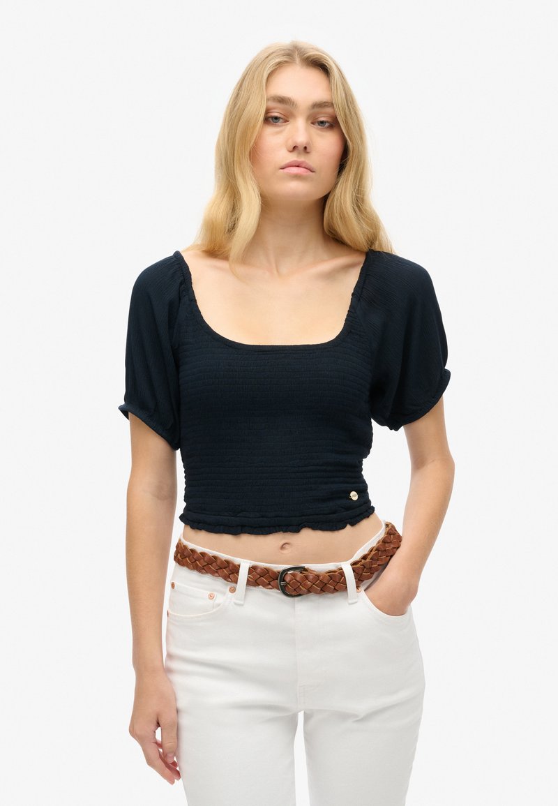 Marineblå cropped top med korte pufærmer, tekstureret smockingdetalje og rund halsudskæring, parret med hvide bukser og et brunt fletbælte.