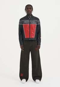 Sort læderjakke med rød frontpanel, sølvdetaljer, lynlås lukning; parret med løse mørke bukser med rød broderi og hvide sneakers.