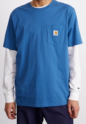 Homme portant une chemise bleue à manches courtes par-dessus une chemise blanche à manches longues avec une poche poitrine et un écusson logo sur la poche.
