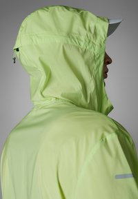 Leichte wasserdichte Jacke in leuchtendem Limettengrün, mit Kapuze, glatter Textur und subtilen reflektierenden Akzenten in der Nähe der Schulter.