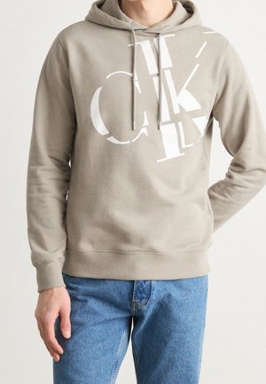 Persoon draagt beige hoodie met witte abstracte letters en een blauwe spijkerbroek, staat met één hand in de zak tegen een effen achtergrond.
