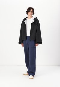 Veste à capuche noire avec doublure grise, poches zippées, portée sur une chemise blanche et un pantalon de survêtement bleu marine. Des baskets beige et rouges complètent la tenue.