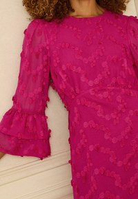 Fuksia värvi kleit läbipaistvate varrukatega, millel on kihilised volangid ja tekstuuriga detailid, kaunistatud samas toonis ringikujuliste dekoratiivsete elementidega.