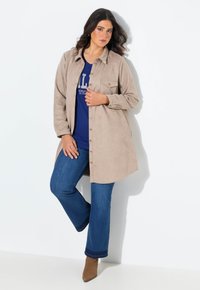 Beige Velourslederhemdjacke mit langen Ärmeln und Knopfverschluss, kombiniert mit einem blauen T-Shirt und ausgestellten blauen Jeans. Braune Stiefeletten.