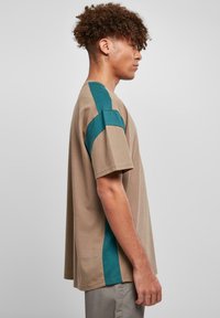 Beige T-Shirt mit kurzen Ärmeln, ausgestattet mit einem tealfarbenen Seitenelement, aus Baumwolle gefertigt, mit lockerer Passform und Rundhalsausschnitt.