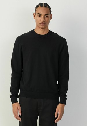 Junger Mann mit geflochtenem Haar, der einen schlichten schwarzen Langarmpullover und schwarze Hose trägt, steht vor einem hellen Hintergrund.