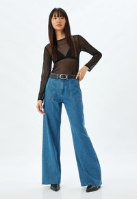 Top negro transparente de manga larga con escote en V, combinado con pantalones de mezclilla anchos de talle alto en azul. Incluye un cinturón negro con hebilla plateada.