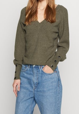 Žena v olivově zeleném žebrovaném svetru s výstřihem do V a knoflíkovými detaily na rukávech a v světle modrých džínách s vysokým pasem.