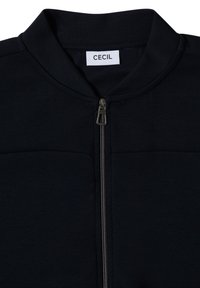 Vêtement zippé noir avec un col montant et une fermeture éclair en argent visible, doté d'une étiquette blanche avec le texte "CECIL" à l'intérieur du col.