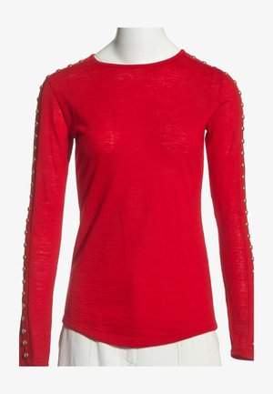 Chemise rouge à manches longues avec col rond et détails de boutons dorés sur chaque manche, portée avec un pantalon blanc sur un mannequin.