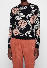 Suéter negro con patrón floral en rosa y blanco. Escote redondo, mangas largas, dobladillo acanalado. Combinado con pantalones de chándal texturizados en naranja.