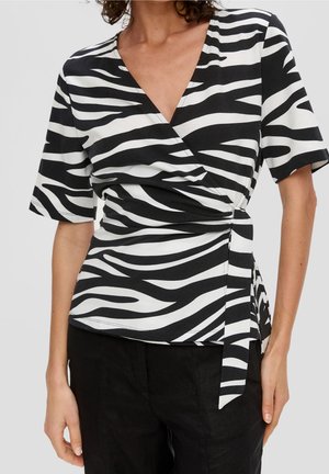 Frau trägt eine schwarz-weiß gestreifte Zebra-Wickelbluse mit kurzen Ärmeln und schwarze Hosen vor einem einfarbigen Hintergrund.