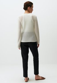 Un pull blanc en maille ouverte à manches longues et col rond, associé à un pantalon noir ajusté ; mannequin de dos. Sandales marron.