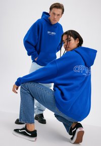 Dva mladí dospělí v modrých mikinách s nápisem „OFF LIMITS“, jeden stojí a druhý dřepí, na jednoduchém bílém pozadí.