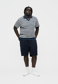 Bărbat în picioare cu mâinile la spate, purtând un tricou cu mâneci scurte cu dungi albastre și albe, pantaloni scurți bleumarin, șosete albe și teniși albi.