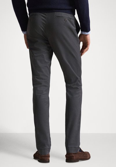Sötétszürke, sima textúrájú chino nadrág, slim fit, hátul zsebekkel, visszafogott fém részletekkel. Barna cipővel viselve.