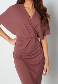 Robe portefeuille mauve avec des manches courtes, un décolleté en V profond, des détails froncés à la taille et une silhouette près du corps. Texture de tissu lisse.