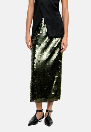 Czarny top bez rękawów z asymetrycznym dołem, w połączeniu z długą, błyszczącą zieloną spódnicą z cekinów oraz czarnymi butami na pasku przy kostce z ostrym noskiem.