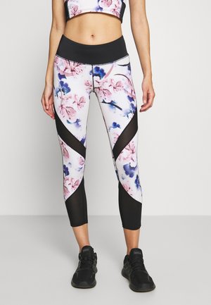 Kvinde iført leggings med blomsterprint og sorte meshpaneler samt en matchende sports-bh, kombineret med sorte Adidas-sneakers.