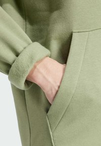 Grön sweatshirt med en mjuk textur, med uppvikt ärm och sidoficka, som framhäver en minimalistisk design.