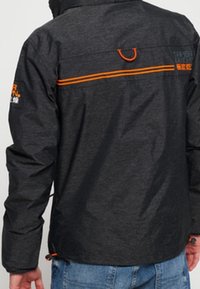 Chaqueta impermeable gris con acentos naranjas, que presenta un cuello alto, dobladillo ajustable y logo en la parte trasera. Textura suave, diseño práctico.
