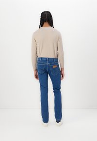 Persona con trenzas largas está de pie frente a una pared blanca, vistiendo una camiseta de manga larga beige, jeans azules con un parche de cuero y zapatillas blancas.