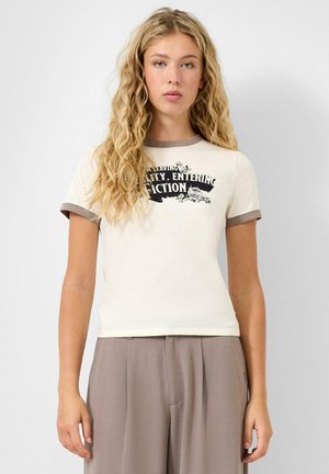 Junge Frau mit langen, lockigen blonden Haaren trägt ein cremefarbenes T-Shirt mit schwarzem Text und taupefarbenem Besatz, kombiniert mit lockeren taupefarbenen Hosen vor weißem Hintergrund.