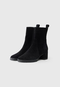 Botines negros de ante con tacón ancho, que cuentan con una cremallera lateral y tiradores en la parte trasera para facilitar el calzado. Textura suave y diseño minimalista.