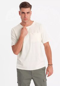 Lys beige kortærmet T-shirt med strukturerede skulderdetaljer og rund hals. Model poserer med hånden på kraven mod en ensfarvet baggrund.
