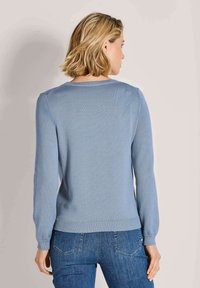 Frau mit kurzen blonden Haaren, die einen hellblauen Langarmpullover und Jeans trägt, mit dem Rücken zur Kamera vor einem schlichten Hintergrund.